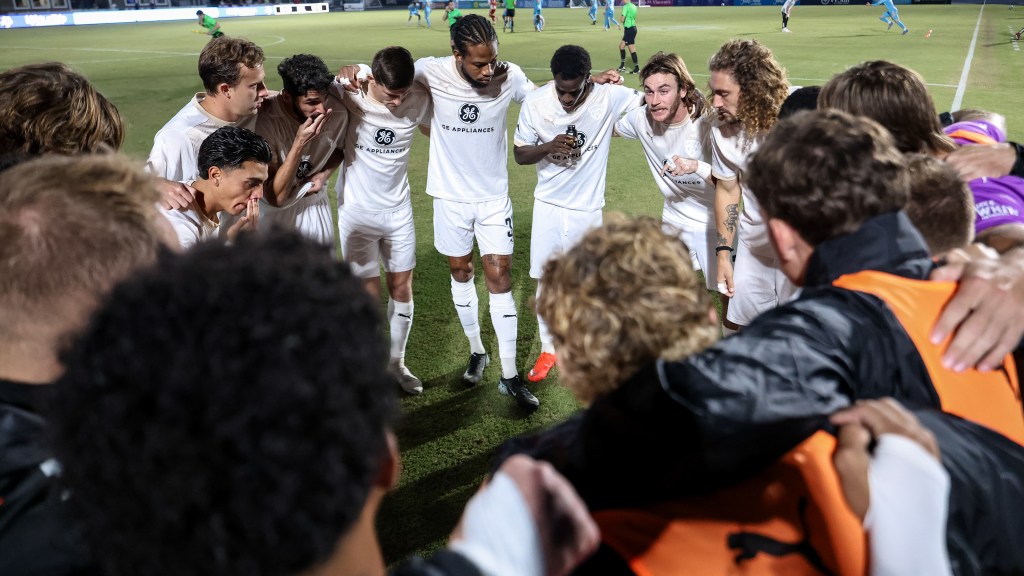 A group of soccer players in white uniforms huddle together on a field at night, embracing and showing camaraderie.